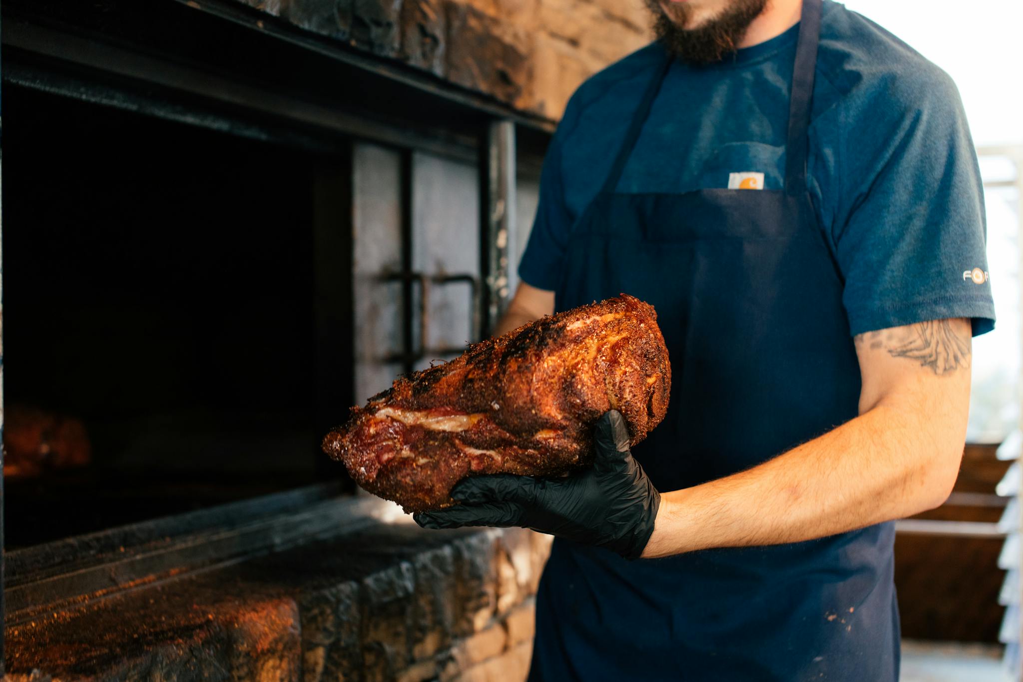 Chef holding seasoned meat with gloves near barbecue pit, cooking outdoors.